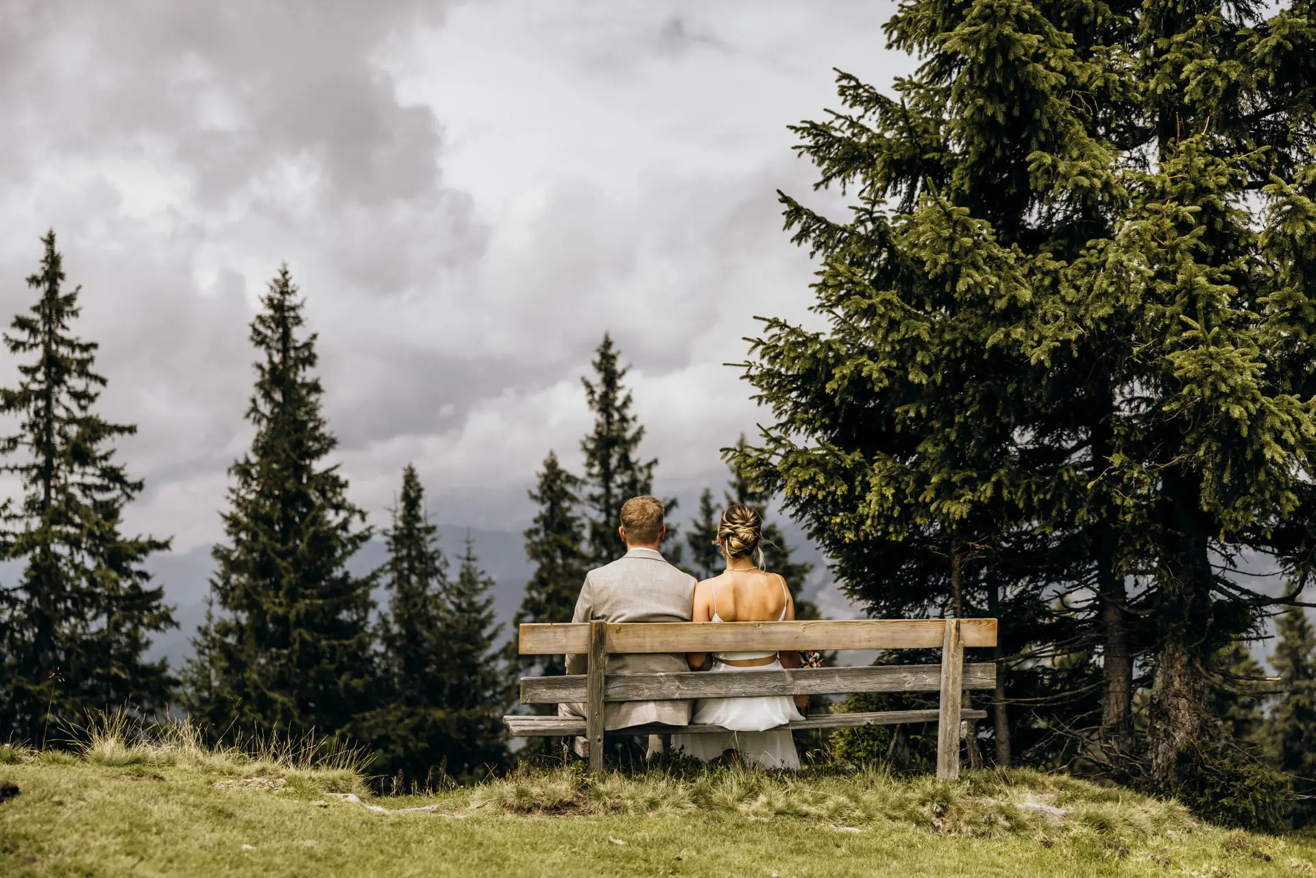 Ein Brautpaar sitzt auf einer Bank bei ihrer intimen Hochzeit in den bergen und schaut ins Tal.