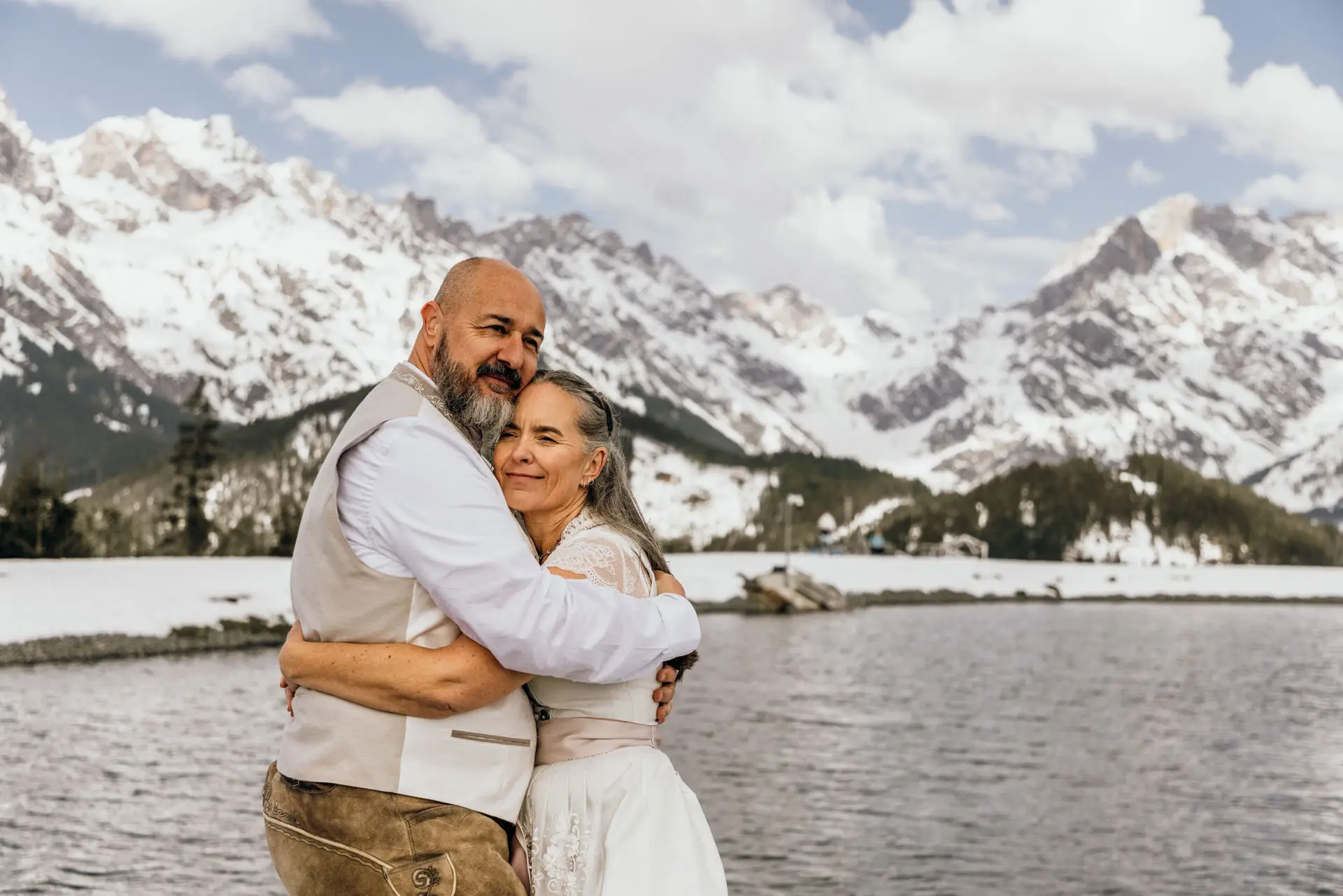 Ein älteres Brautpaar umarmt sich liebevoll vor einer atemberaubenden Kulisse aus schneebedeckten Bergen und einem ruhigen See. Die intime Atmosphäre spiegelt die Schönheit einer Hochzeit ohne Gäste wider – ein Moment der puren Verbundenheit und Zweisamkeit in der Natur.